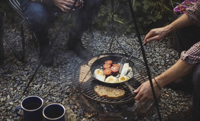 A campfire scene with people sitting around it (just visible). Cooking on a barbecue over the fire is some toast, frying eggs in a pan and fried tomat